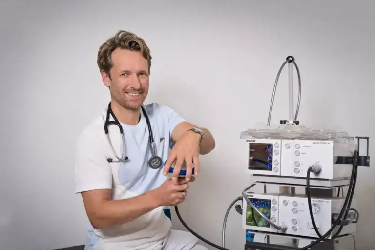 Ein lächelnder Arzt in weißer Kleidung mit Stethoskop sitzt neben einem modernen medizinischen Gerät zur Behandlung, vor einem neutralen Hintergrund, Stoßwellengerät, Stoßwellentherapie, Stoßwellenbehandlung, Stoßwellentherapie Kosten