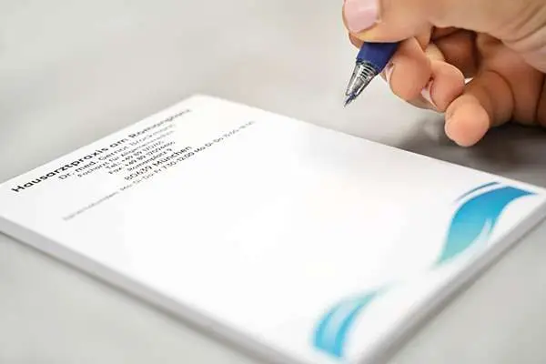 Ein Hand hält einen Stift über ein Rezeptpapier mit blauer Wellenverzierung, bereit zum Schreiben. OP-Vorbereitung